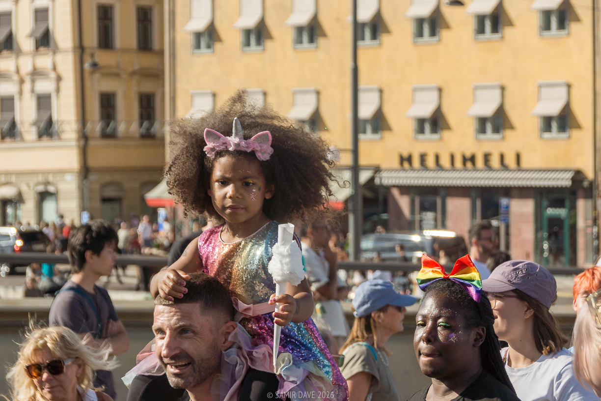 A little child dressed as unicorn sitting on the shoulders of an adult in the afternoon heat at Stockholm Pride Festival 2026.
